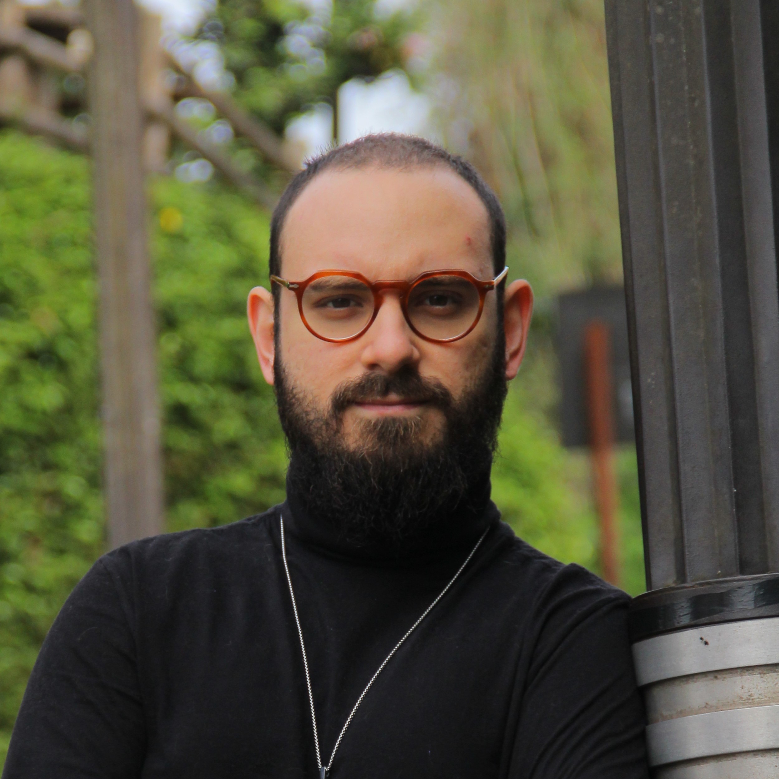 A man with a neatly trimmed beard and short hair, wearing round, brown glasses and a black turtleneck sweater. He is standing outdoors next to a tall, dark-colored pole, with a blurred green leafy background. The man has a calm and confident expression, with his arms crossed lightly in front of him. He is also wearing a thin silver necklace.